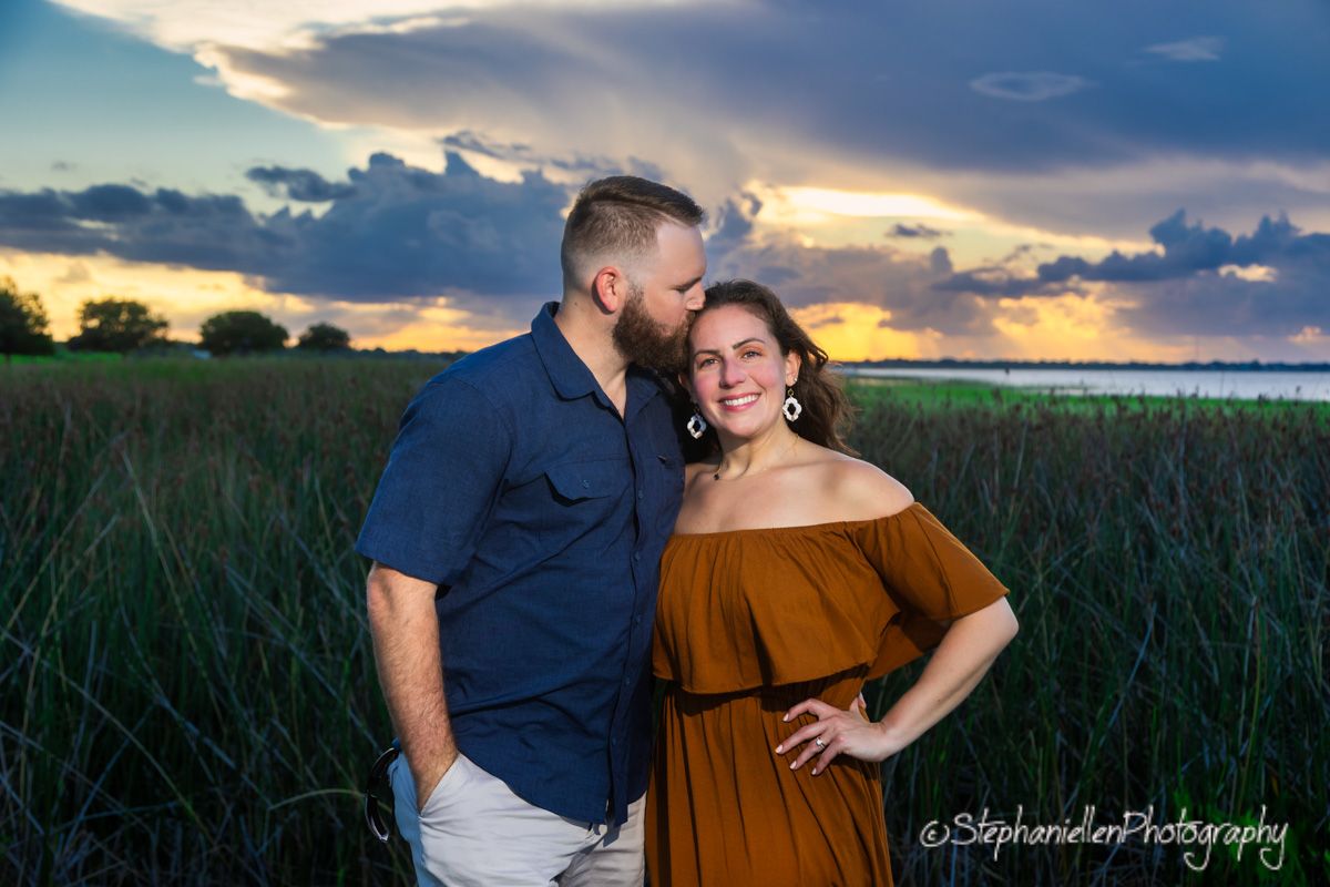Couple embracing in a field at sunset with dramatic clouds, woman in rust dress and man in navy shirt.