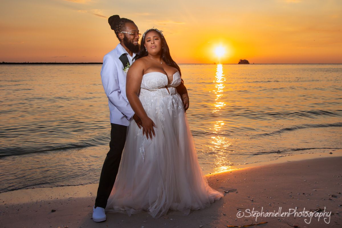 Bride in flowing gown and groom in white suit posing on beach during golden sunset.