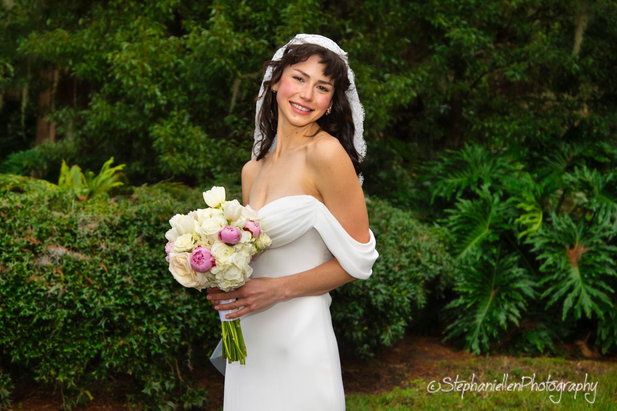 Smiling bride in white gown holds elegant bouquet in lush garden setting.