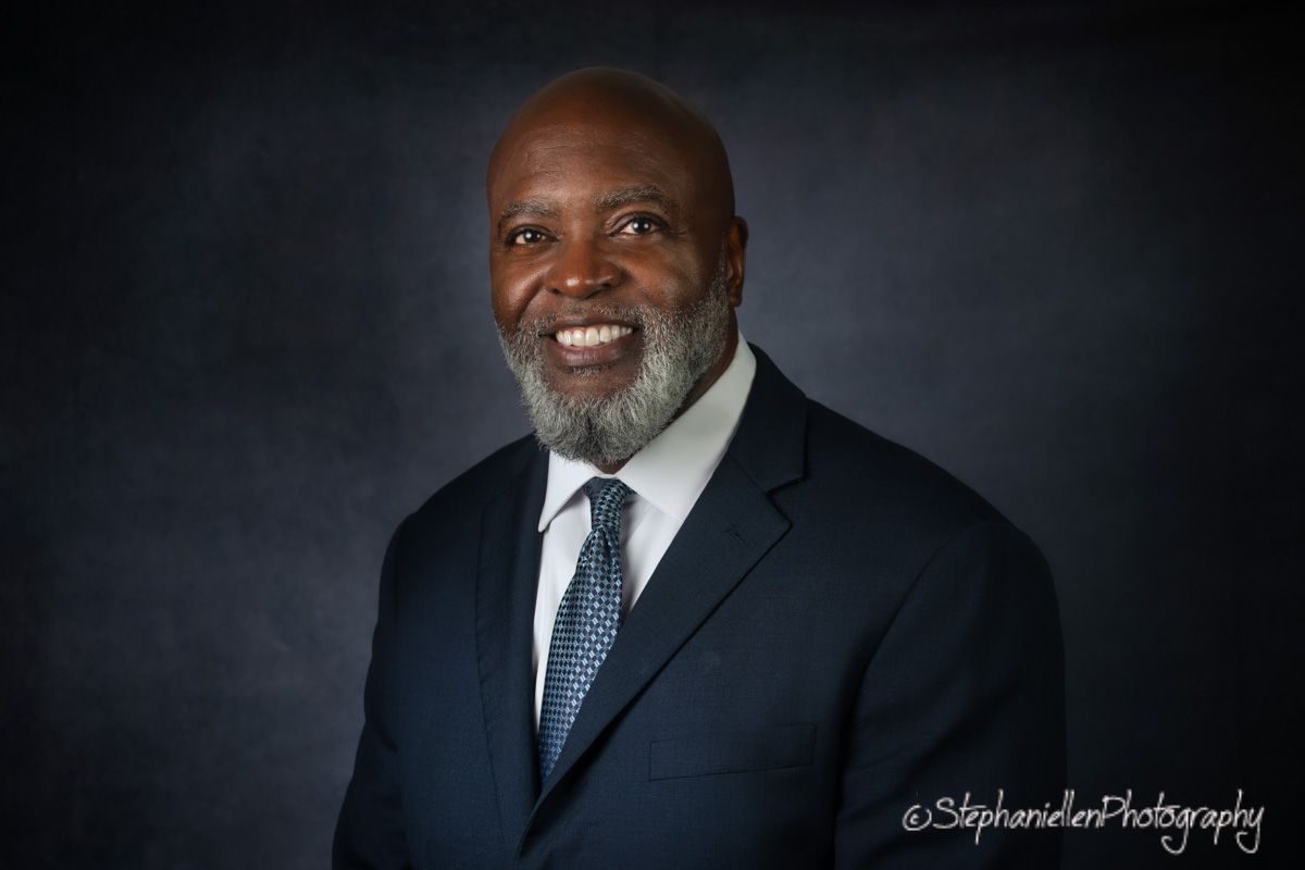 Professional headshot of a smiling man in navy suit and tie against dark background.