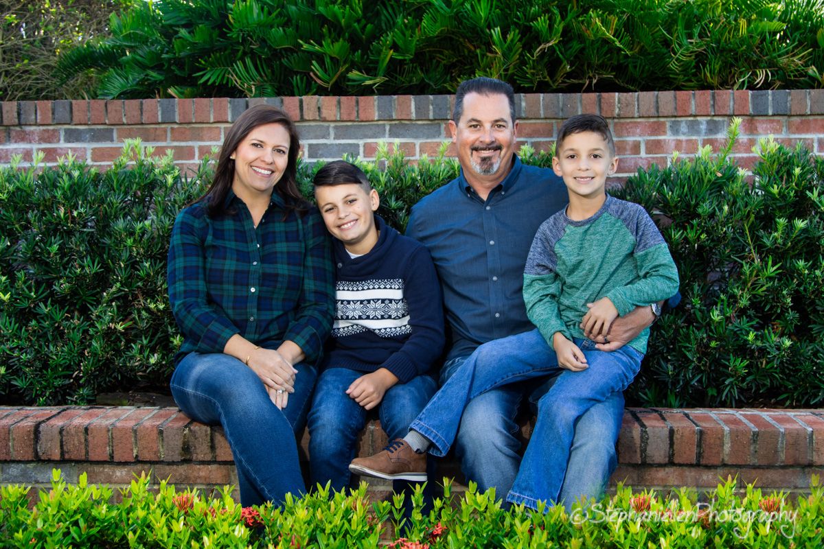 Family of four sitting on brick steps smiling outdoors with green bushes background.