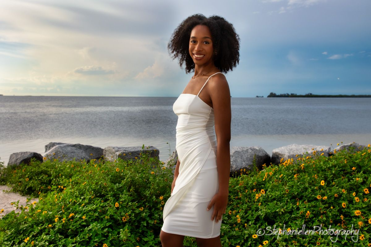 Young woman in white dress standing near the waterfront with wildflowers and rocks in background.
