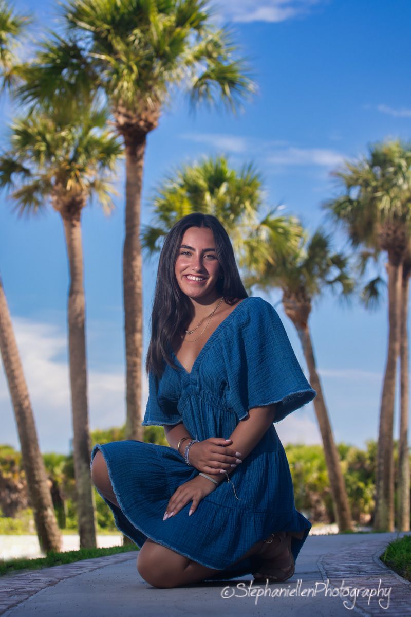 Young woman in blue dress sitting cross-legged outdoors with tall palm trees against blue sky.