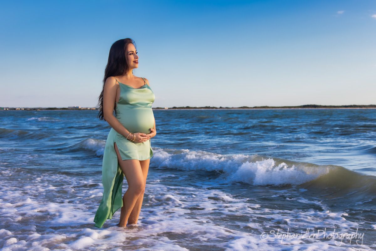 Pregnant woman in a flowing mint green dress standing in ocean waves during a maternity photoshoot.