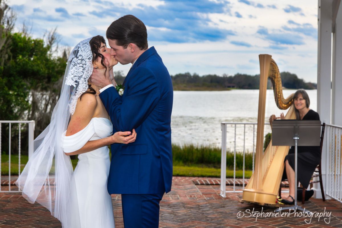 Bride and groom share first kiss with harpist performing by waterfront.