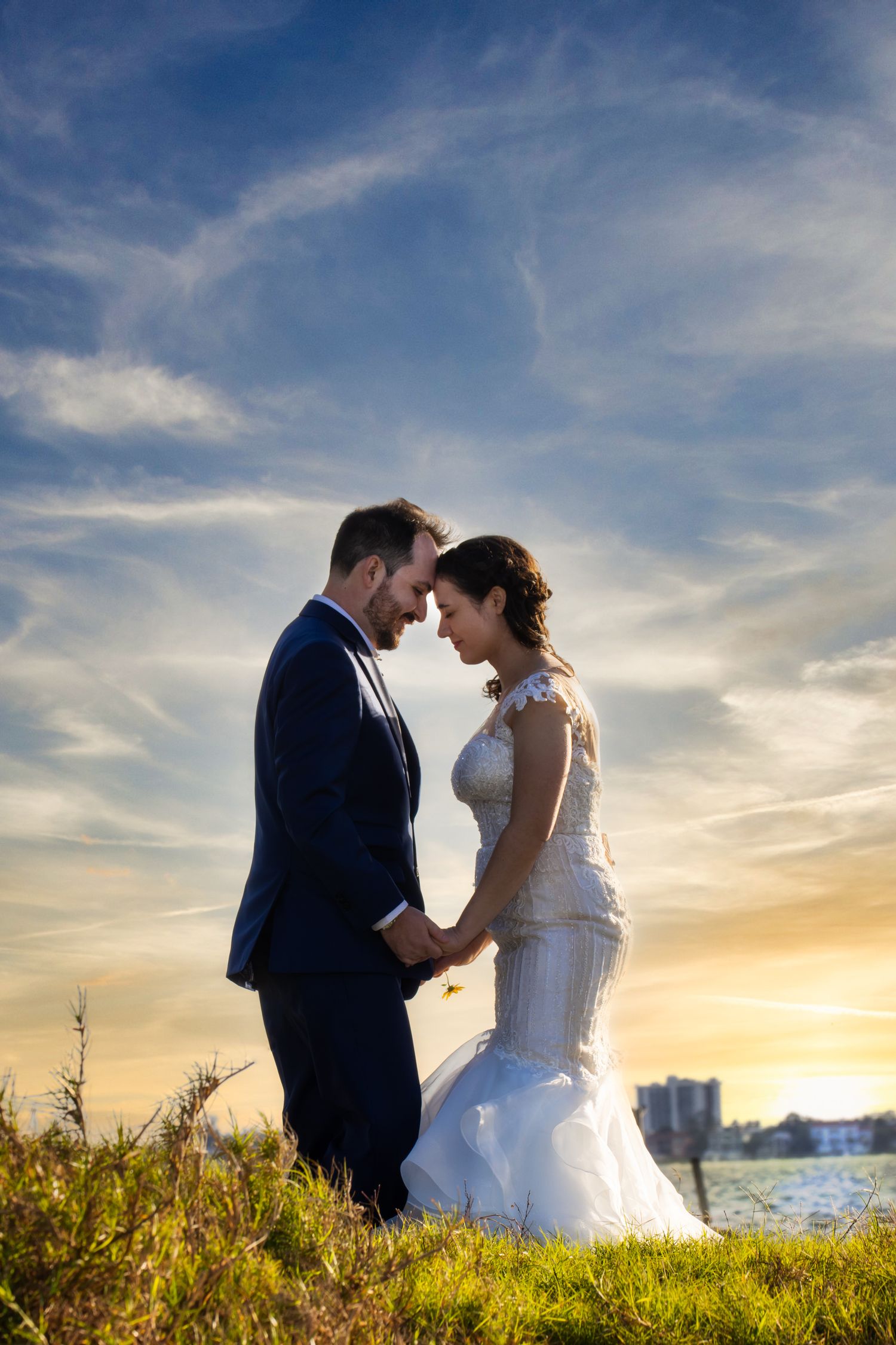 Bride and groom hold hands facing each other at sunset near a waterfront in a romantic pose.