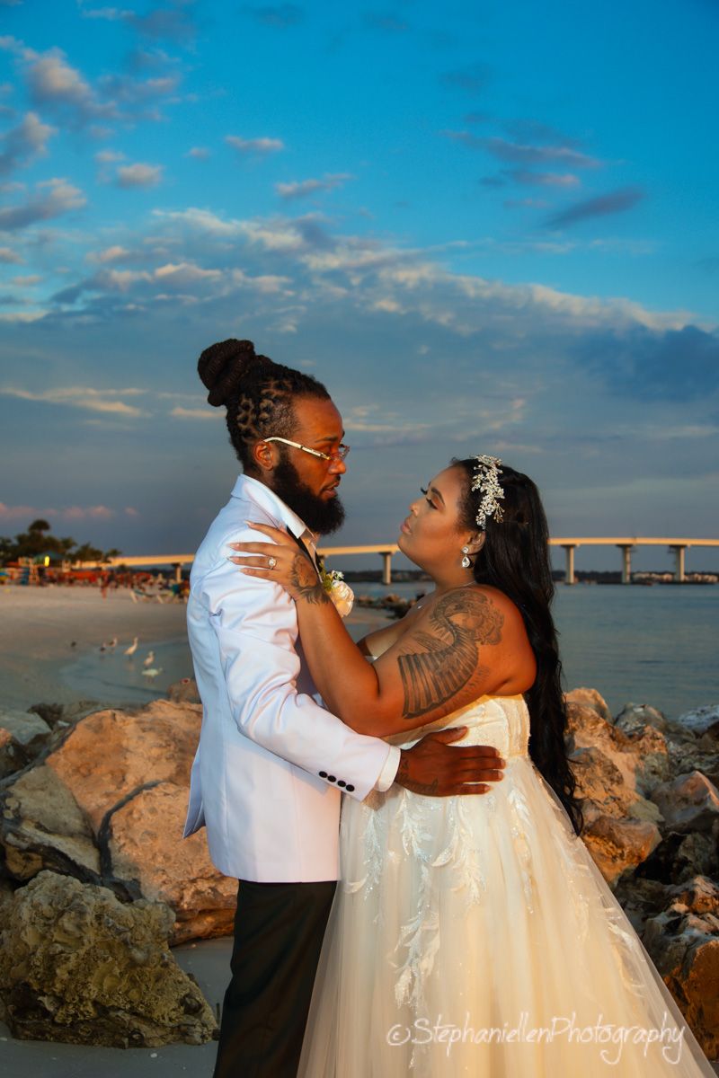 Bride and groom embracing on rocky beach at sunset with bridge in background.