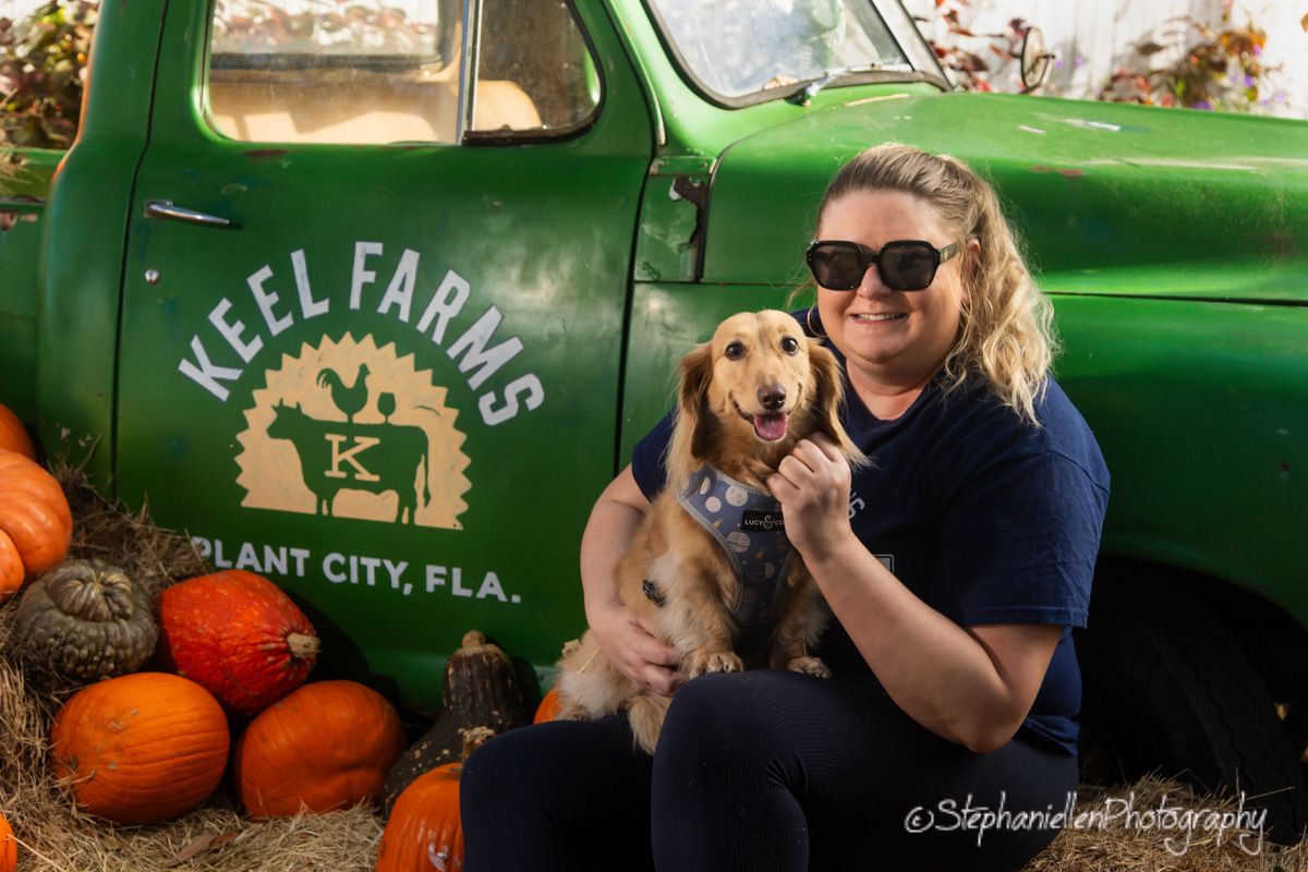 Woman with sunglasses hugging golden dog beside Keel Farms green truck surrounded by colorful pumpkins in Plant City FL.