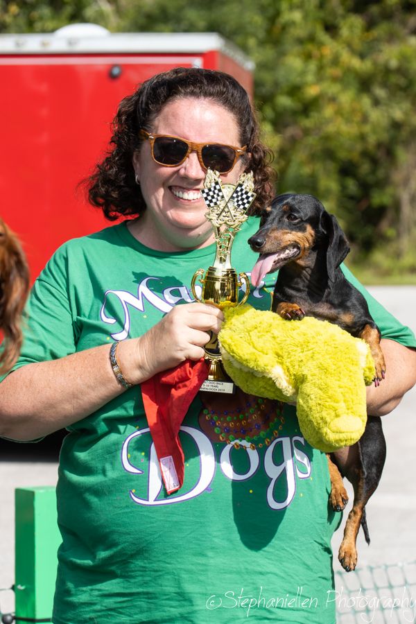 Smiling woman in green Dogs shirt holds a dachshund in yellow costume and a racing trophy outdoors.