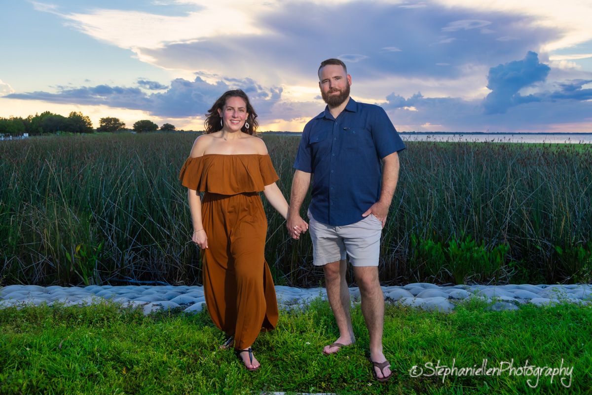 Couple holds hands standing before a marsh and dramatic sunset sky in an outdoor portrait.