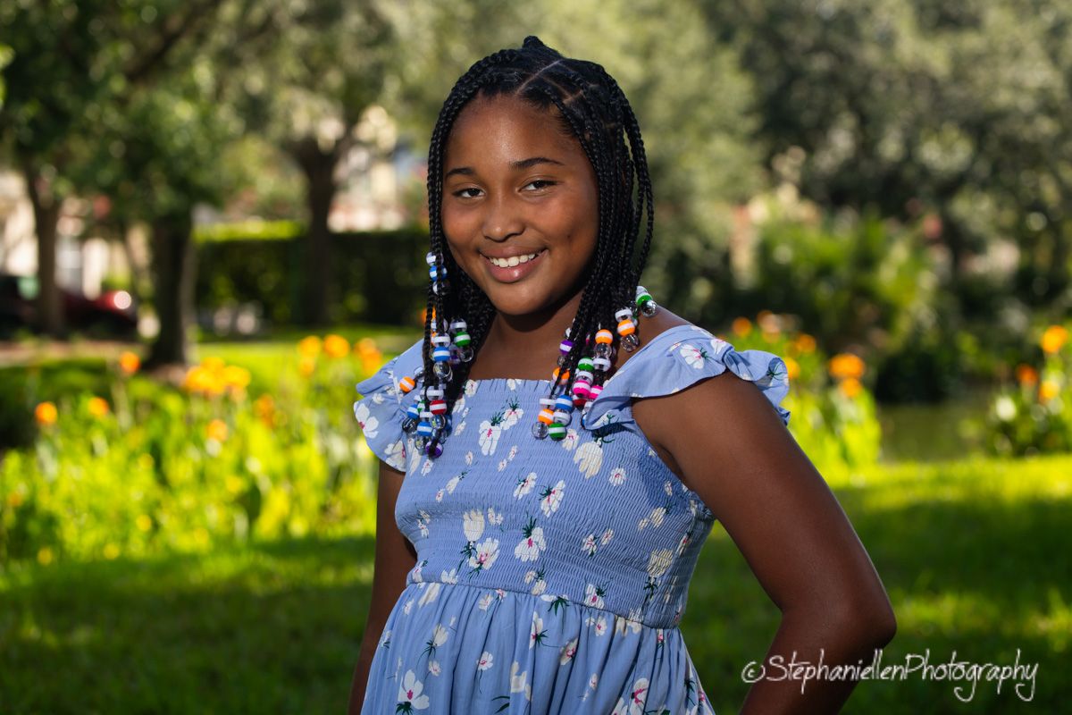 Young girl with colorful beaded braids smiles in a blue floral dress outdoors in a lush green park setting.
