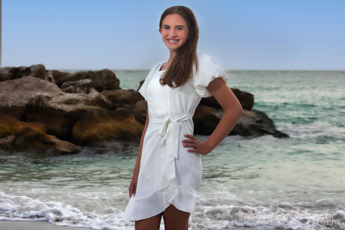 Young woman in white dress posing on a sunny beach with rocky shoreline and turquoise ocean waves behind her.