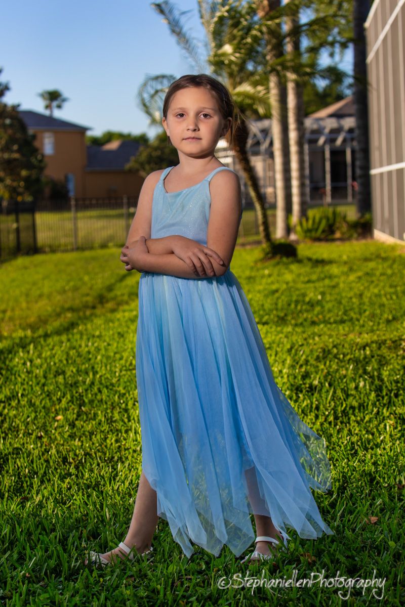 Young girl in a blue tulle dress stands with arms crossed on green grass in a sunny backyard setting.