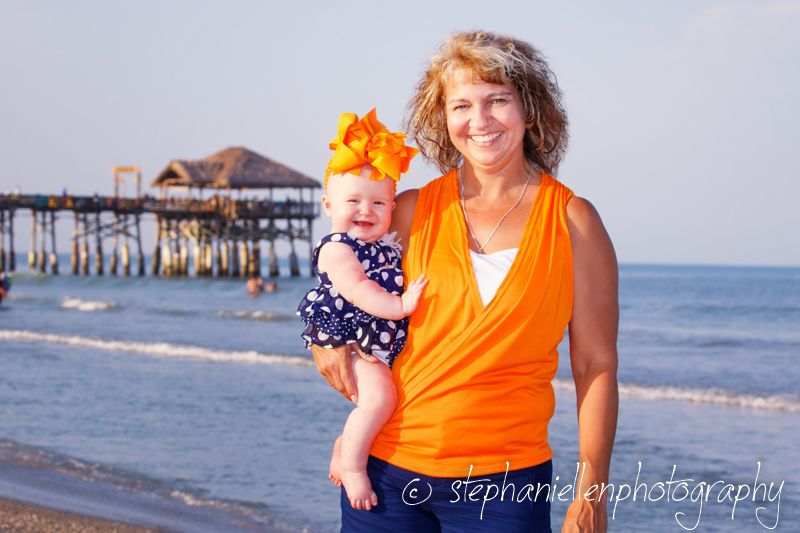 Woman in orange top holds baby girl with orange bow on beach near pier.