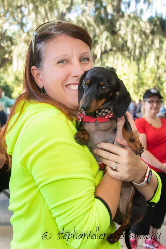 Woman in neon yellow shirt smiling while holding a black and tan Dachshund with a red collar outdoors.