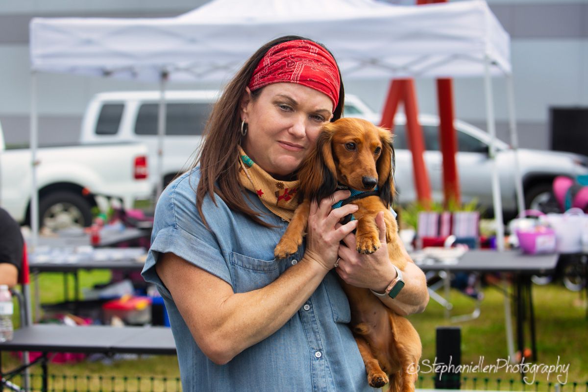Woman in red bandana holds a golden dachshund at an outdoor event, smiling warmly at a vendor fair.