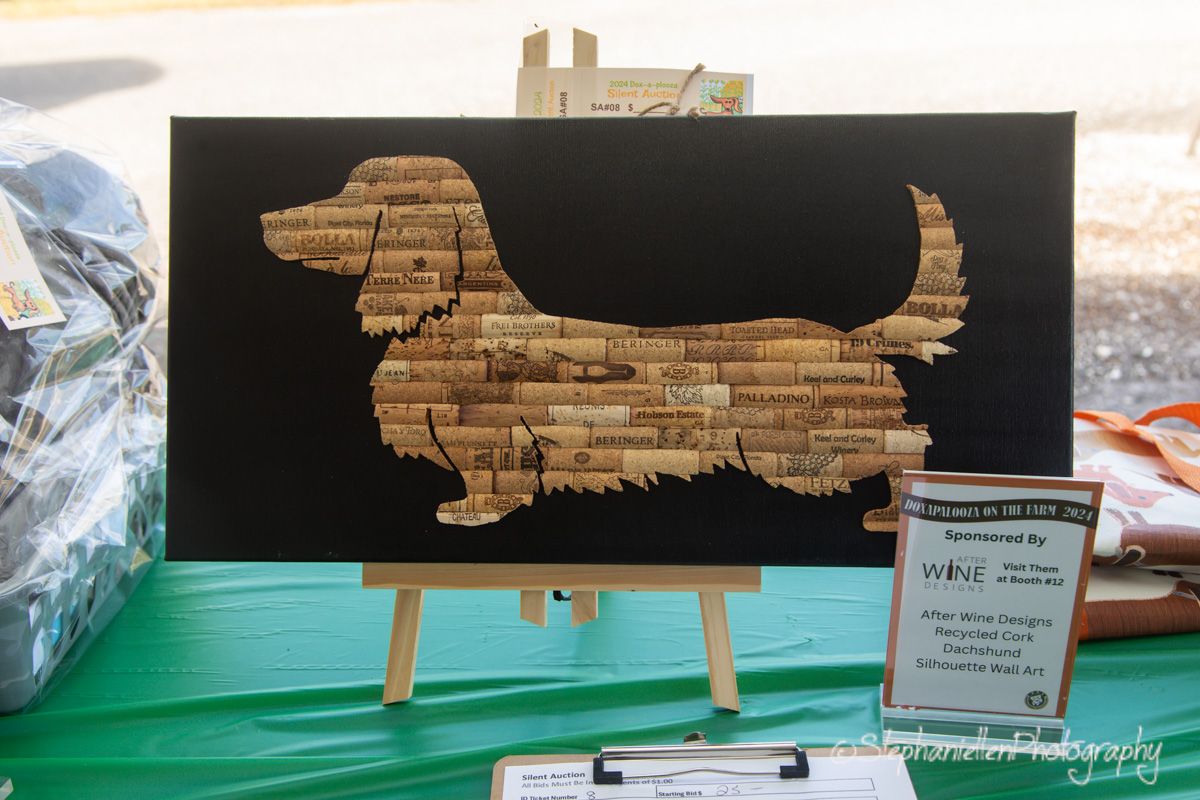 Cork art dachshund dog silhouette on black canvas displayed on wooden easel at outdoor wine event.