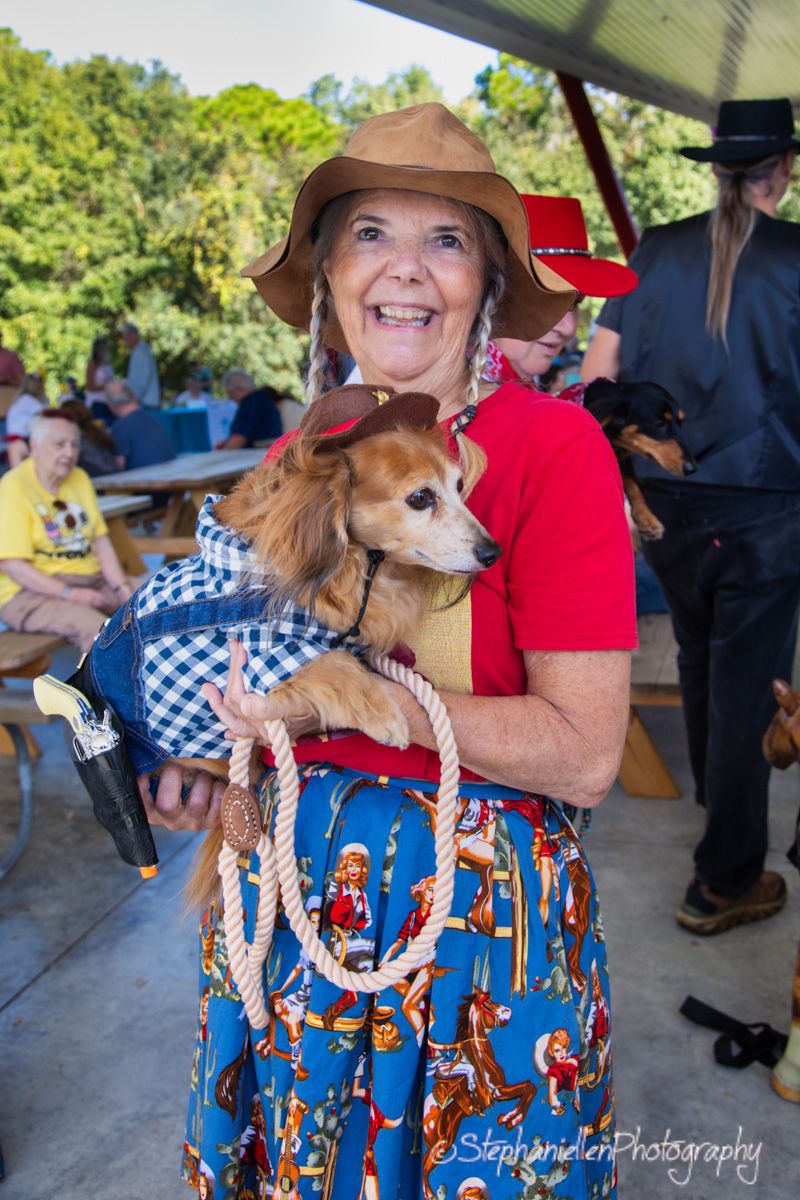 Smiling woman in cowboy hat and western dress holds a dachshund in a gingham outfit at an outdoor event.
