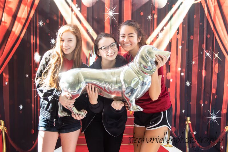Three girls smiling and posing with a silver dog balloon sculpture in front of a red carpet backdrop.