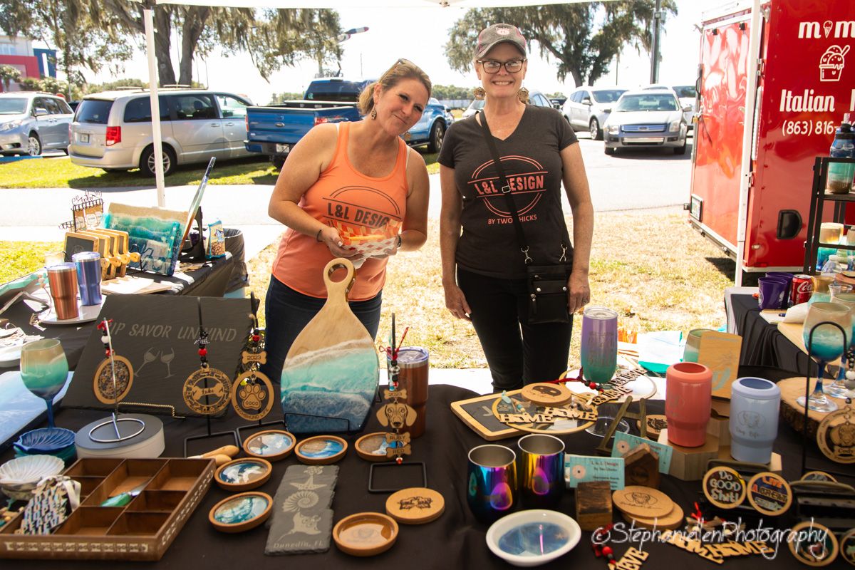 Two women smile behind a craft vendor table displaying handmade wooden art, coasters, and colorful goods at an outdoor market.