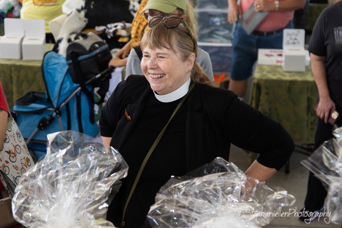 Smiling woman in clergy collar holding wrapped gift baskets at an outdoor market event.