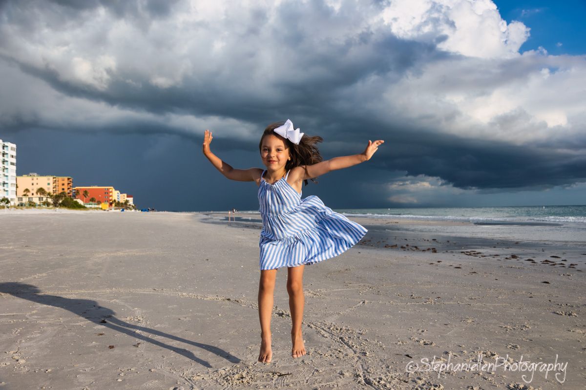 Young girl in a blue striped dress jumping joyfully on a beach with stormy skies behind her.