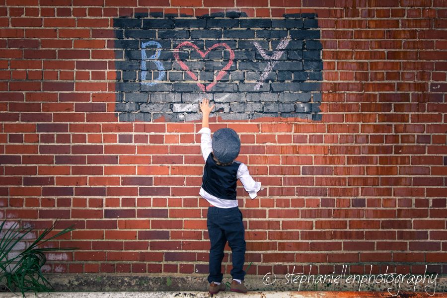 A toddler in a vest reaches up to touch graffiti art on a red brick wall featuring the word BOY with a heart.