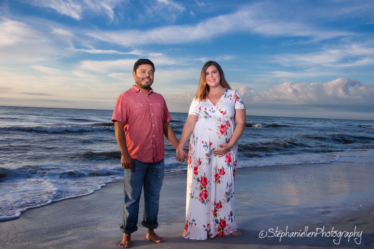 Pregnant couple posing on a sandy beach at sunset with blue sky and ocean waves behind them.