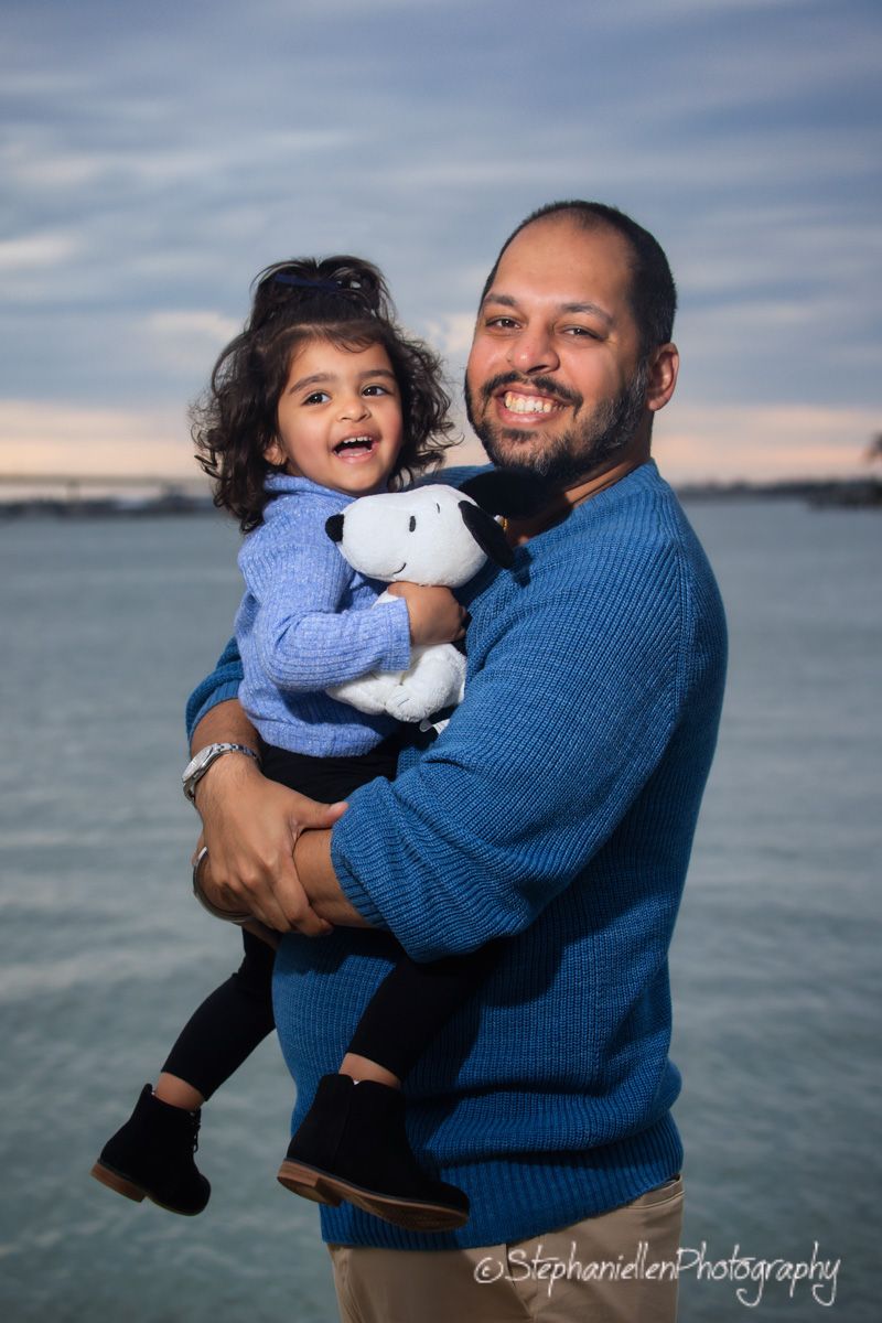 A smiling father holds his laughing toddler clutching a Snoopy plush toy near the waterfront at dusk.
