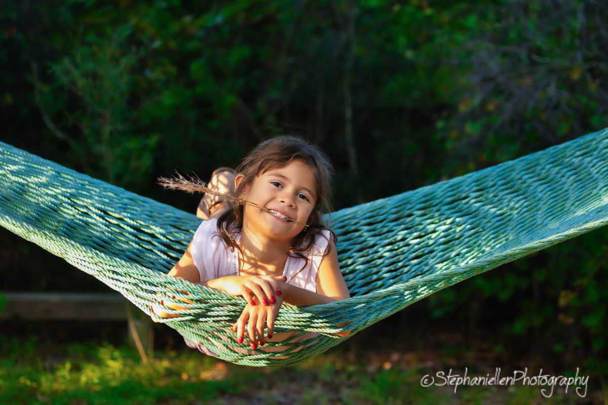 A smiling young girl relaxing in a teal hammock outdoors, surrounded by lush green trees in warm sunlight.