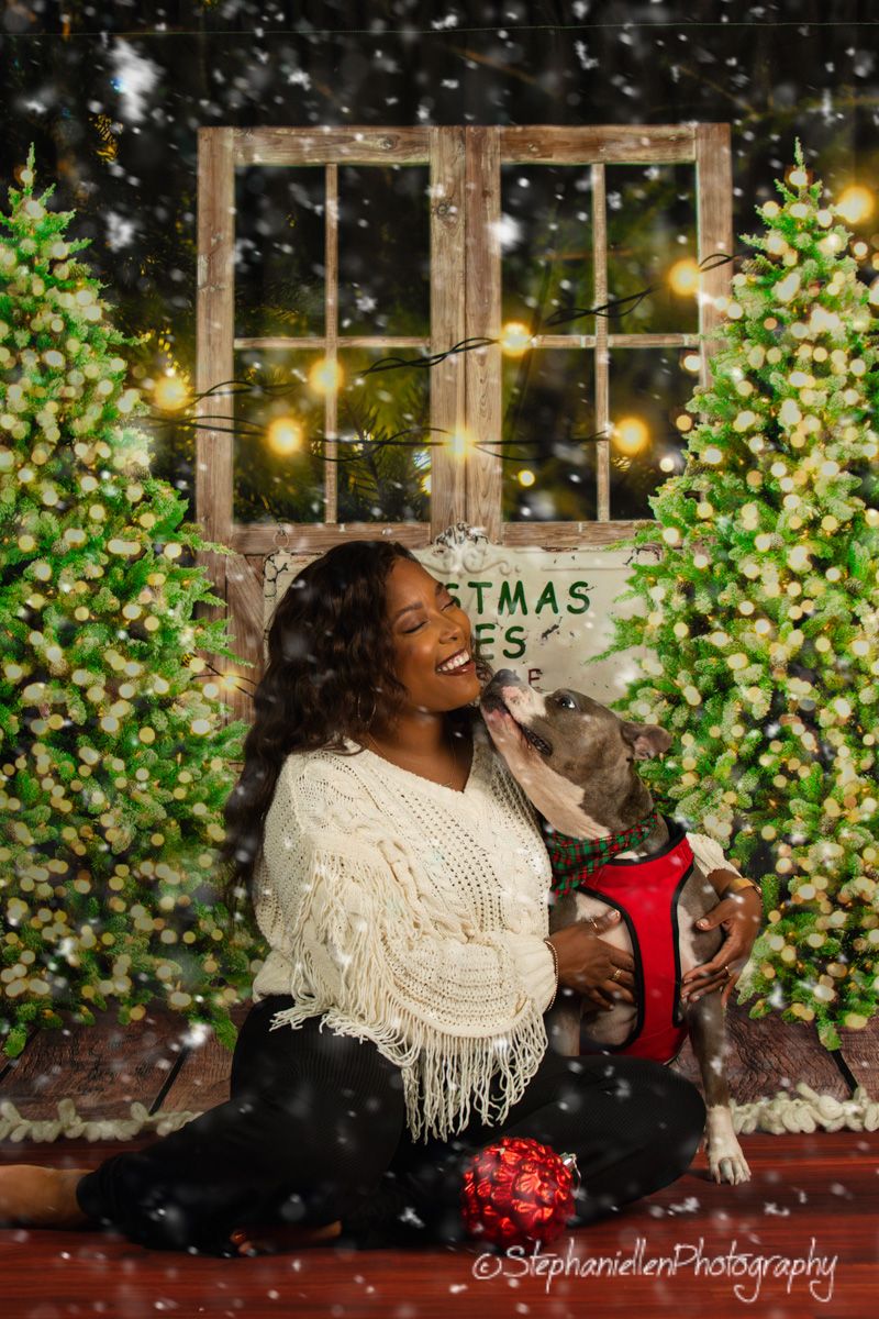 Woman laughing while holding a small dog in a red harness beside snow-dusted Christmas trees.