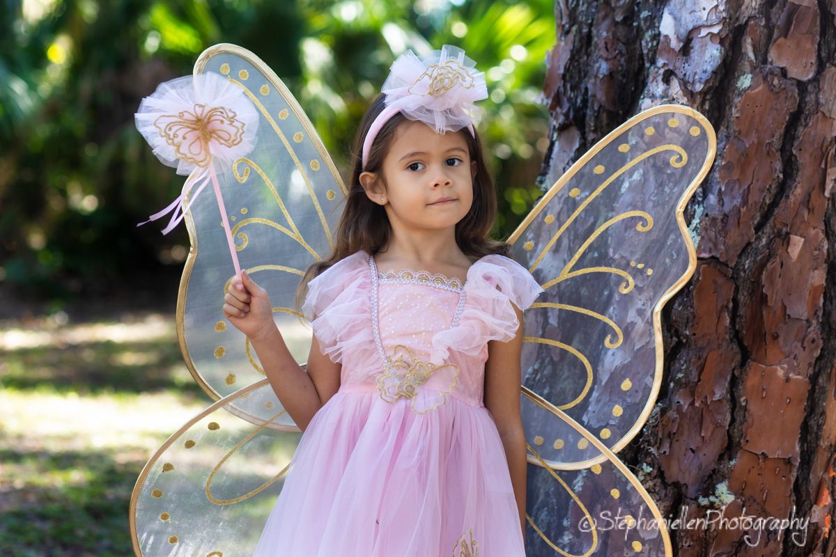Adorable little girl dressed in a pink fairy costume with butterfly wings holding a wand outdoors.