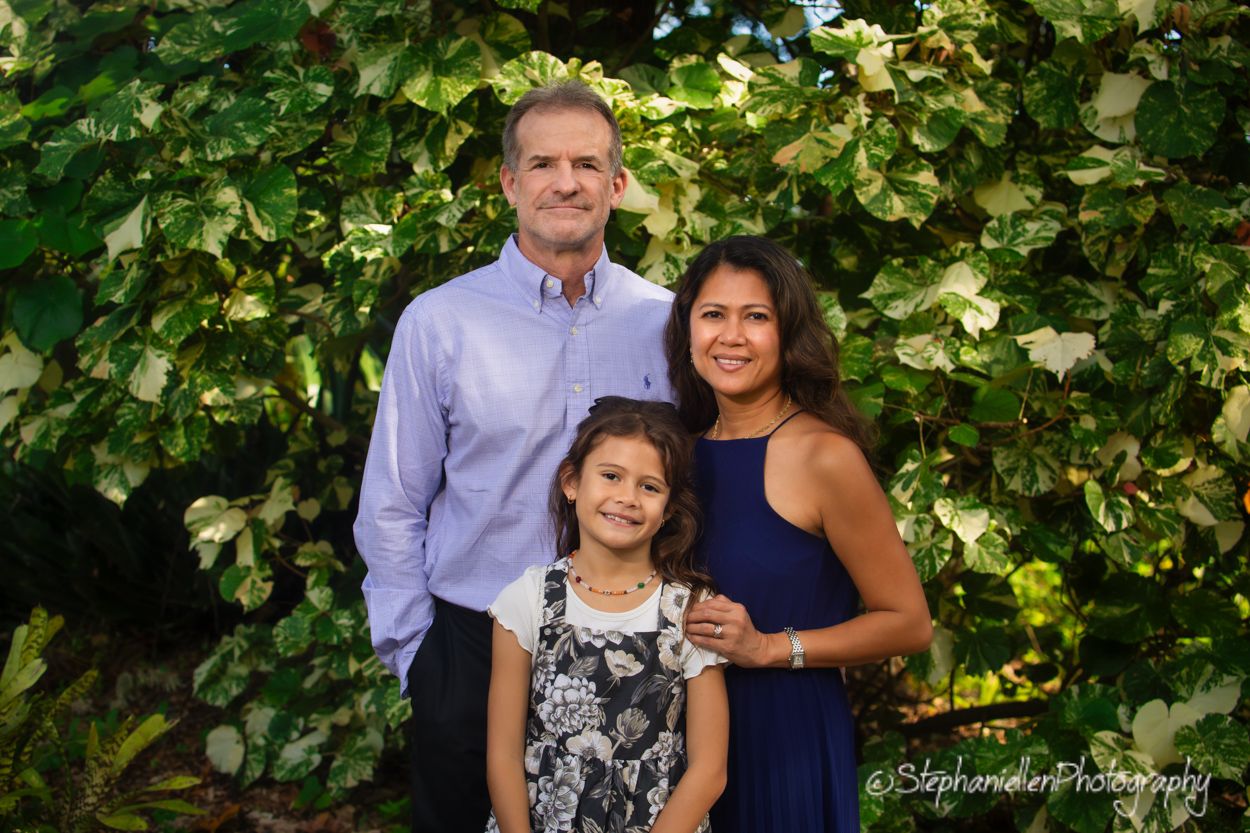 Family of three portrait with man in blue shirt, woman in navy dress, and young girl outdoors.