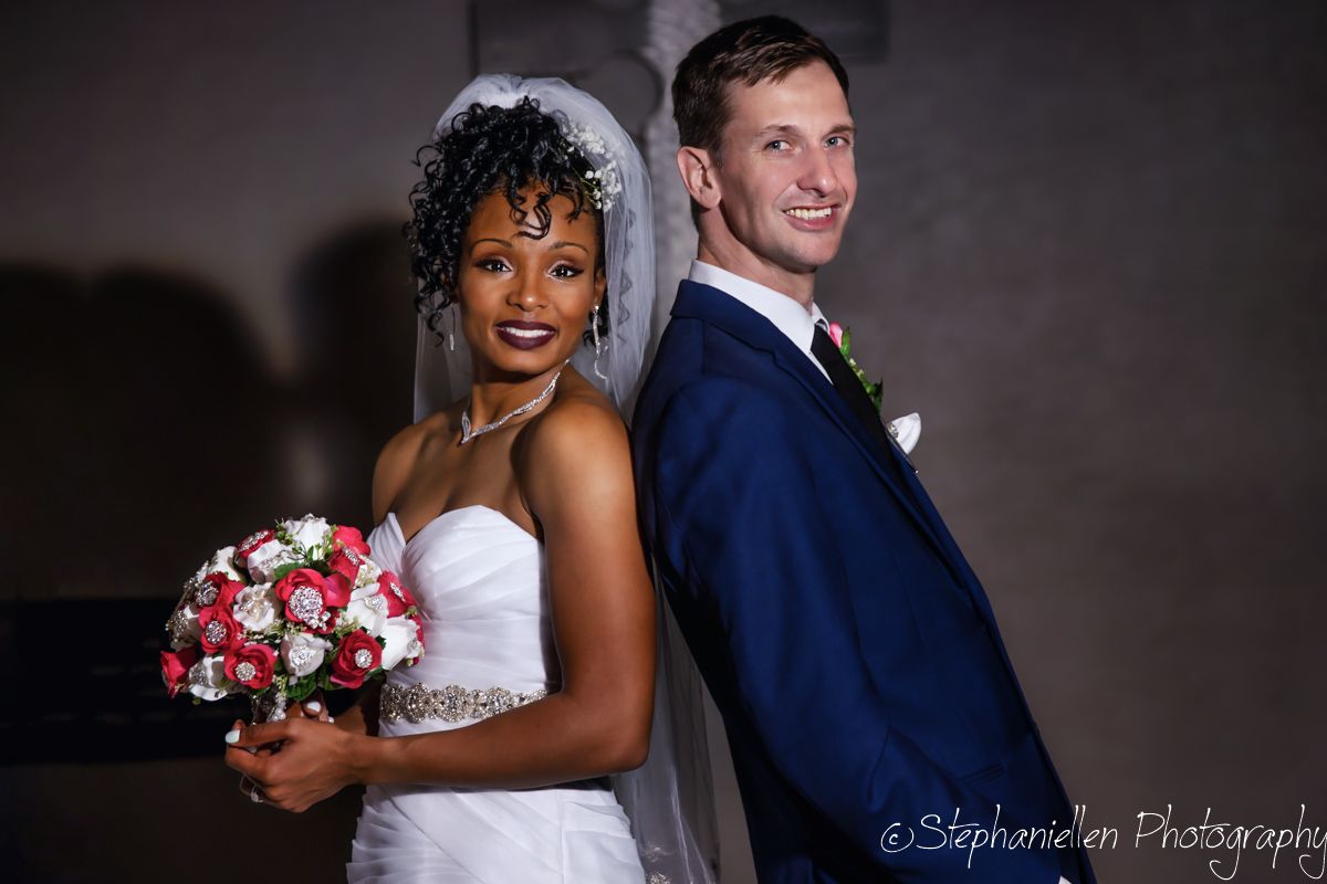Smiling interracial bride and groom posing back-to-back with a bridal bouquet at their wedding venue.