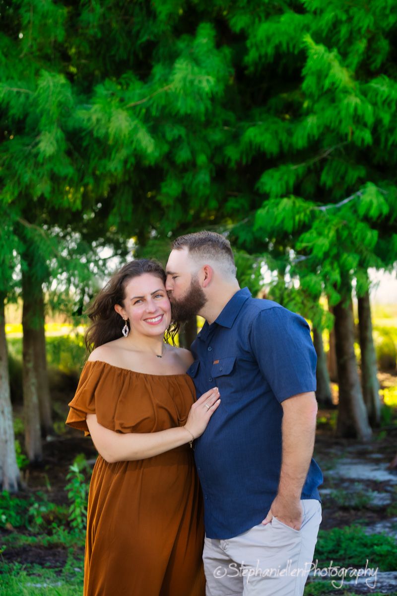 Couple poses outdoors, woman in rust off-shoulder dress smiles as bearded man in navy shirt kisses her cheek near green trees.