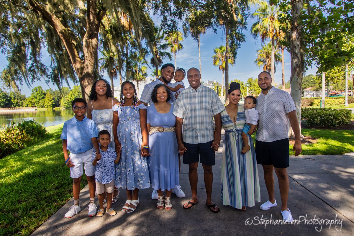 A large multigenerational Black family poses together outdoors near a scenic pond surrounded by lush trees.