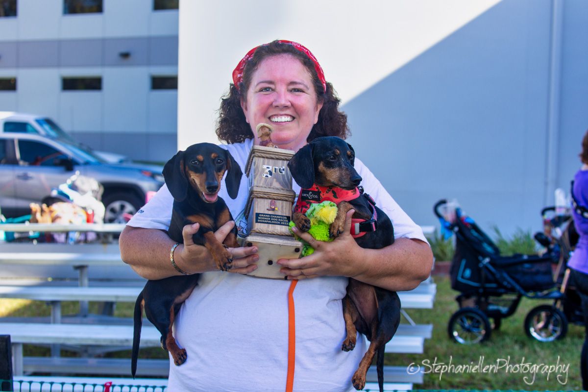 Smiling woman holds two dachshund dogs while standing outdoors at a community event.