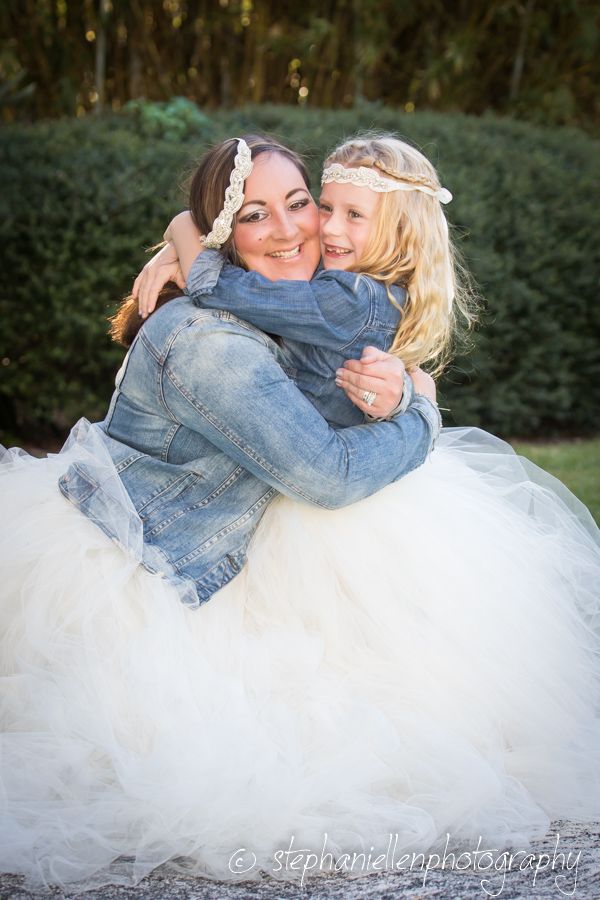 Bride in white tulle skirt and denim jacket hugging a young blonde flower girl outdoors.