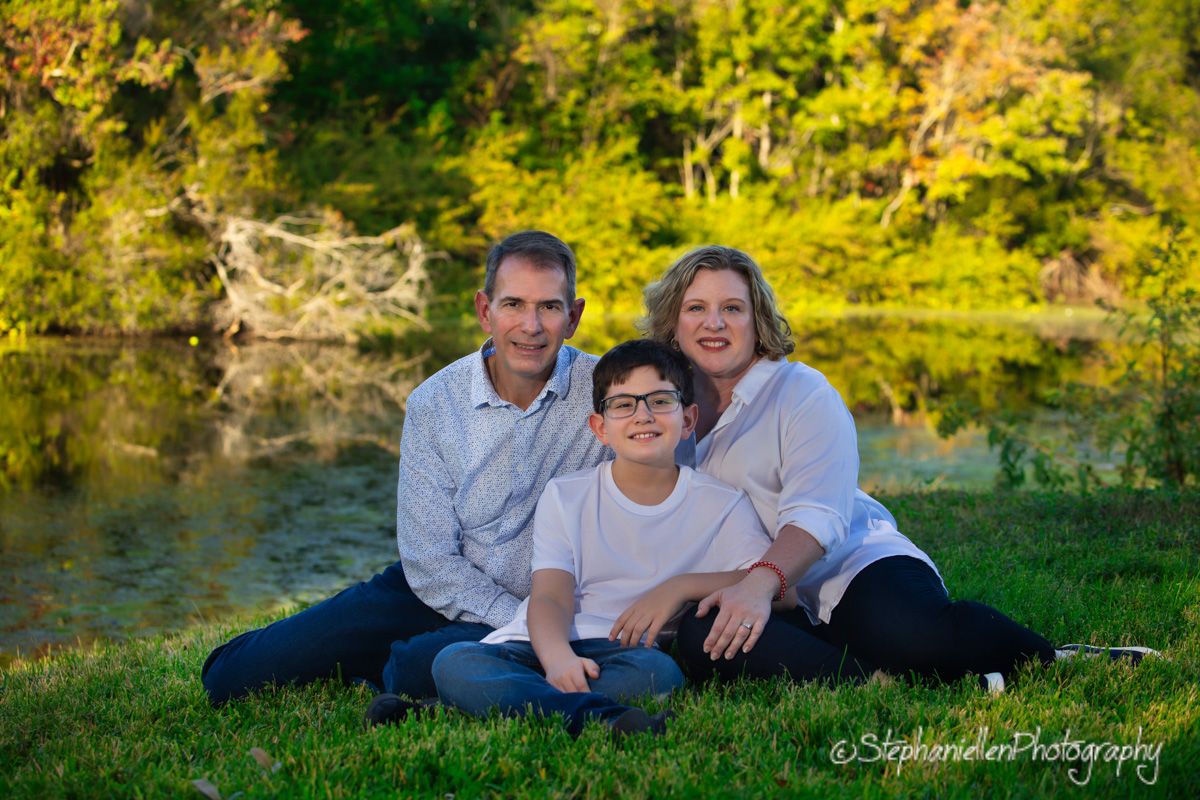 Family of three smiling outdoors by a pond, sitting on green grass surrounded by lush trees in warm golden light.