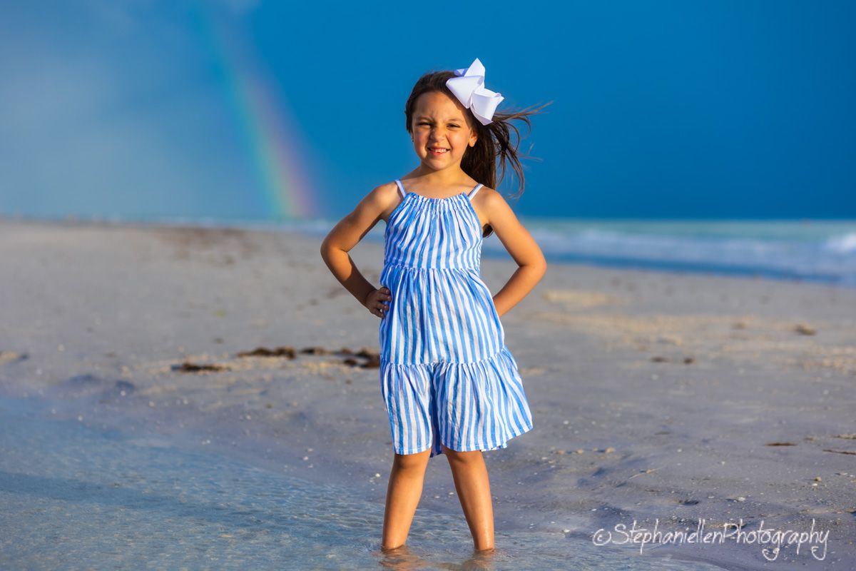 Young girl in blue striped dress posing on beach with rainbow behind her.