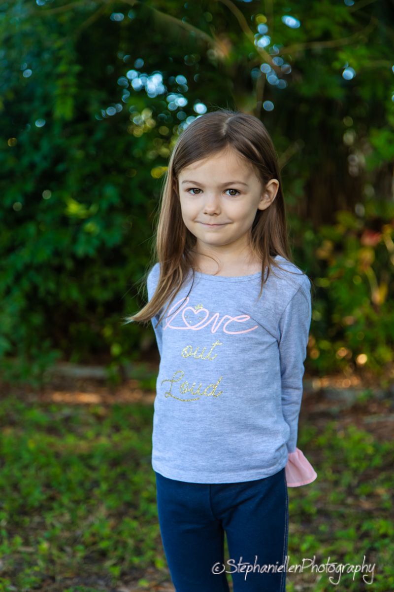 Young girl in gray long-sleeve top and navy pants smiles outdoors, surrounded by lush green trees.
