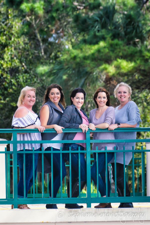 Five women smiling and leaning on a teal railing outdoors, surrounded by lush green trees in a park setting.
