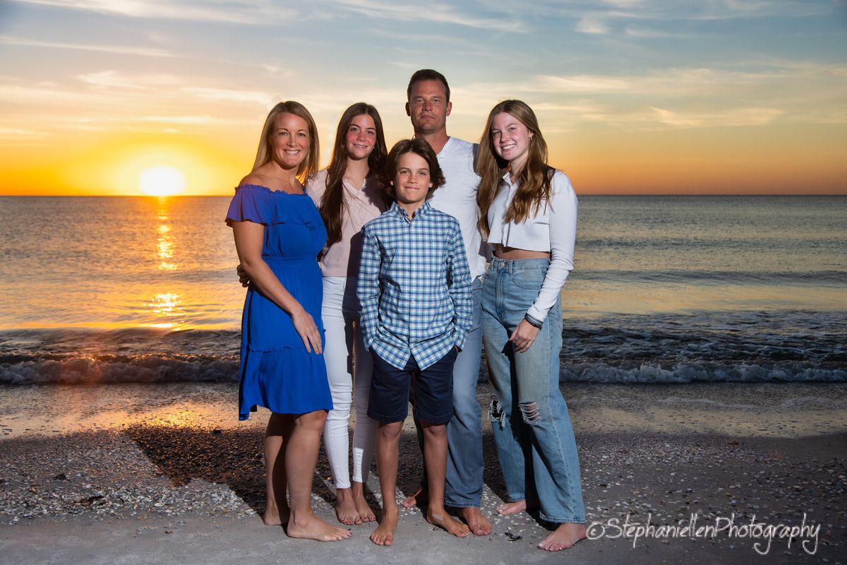 Family of five poses on a sandy beach at sunset with golden sky and ocean waves in the background.