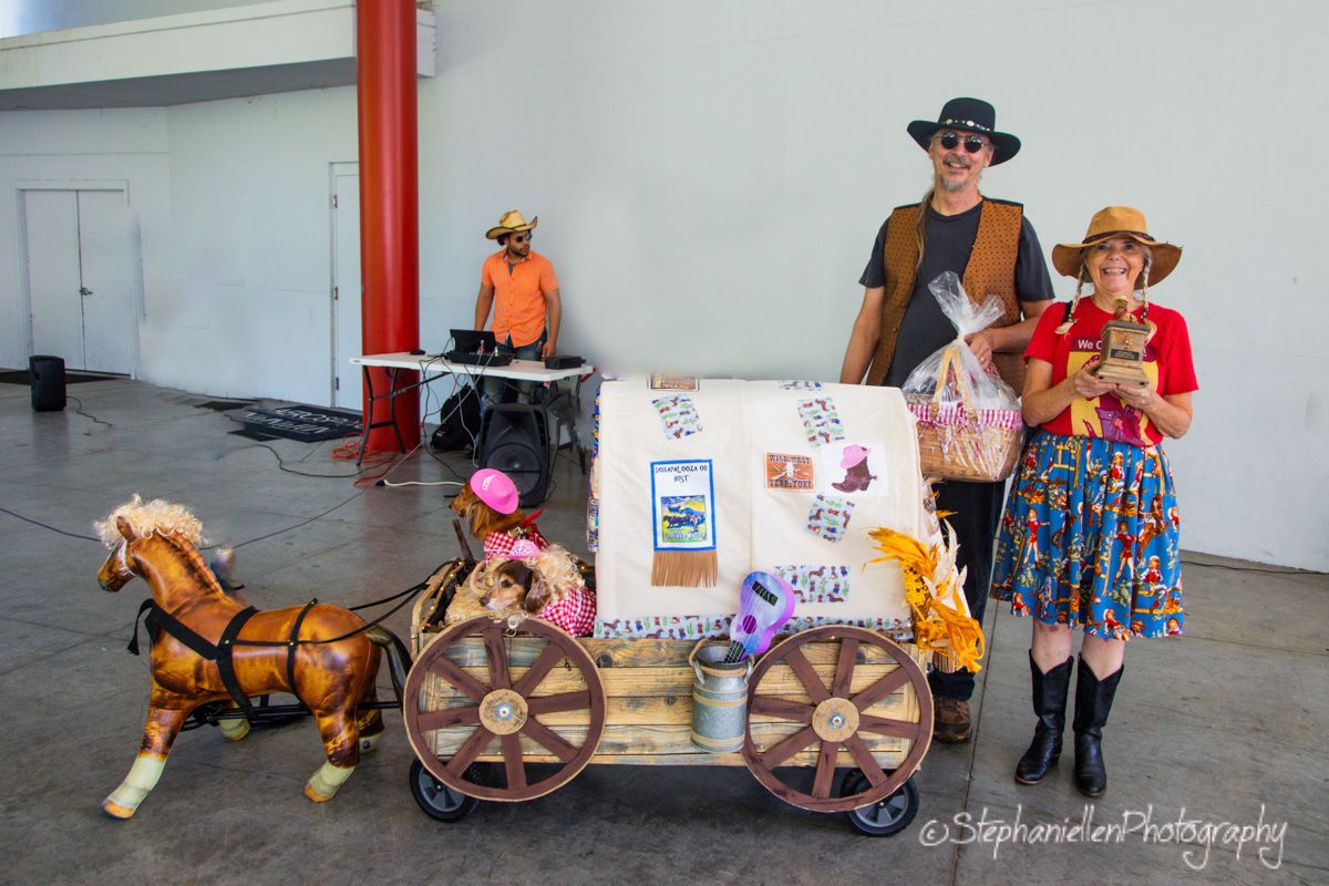 Two people in cowboy costumes pose with a decorated wagon and toy horse at an indoor event.
