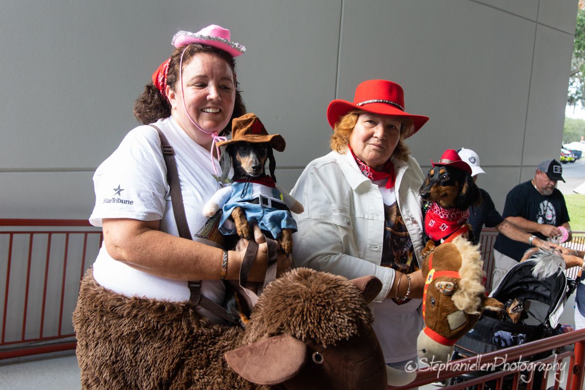 Two women in costumes hold dressed-up dogs at an outdoor pet parade event, wearing fun hats and festive outfits.
