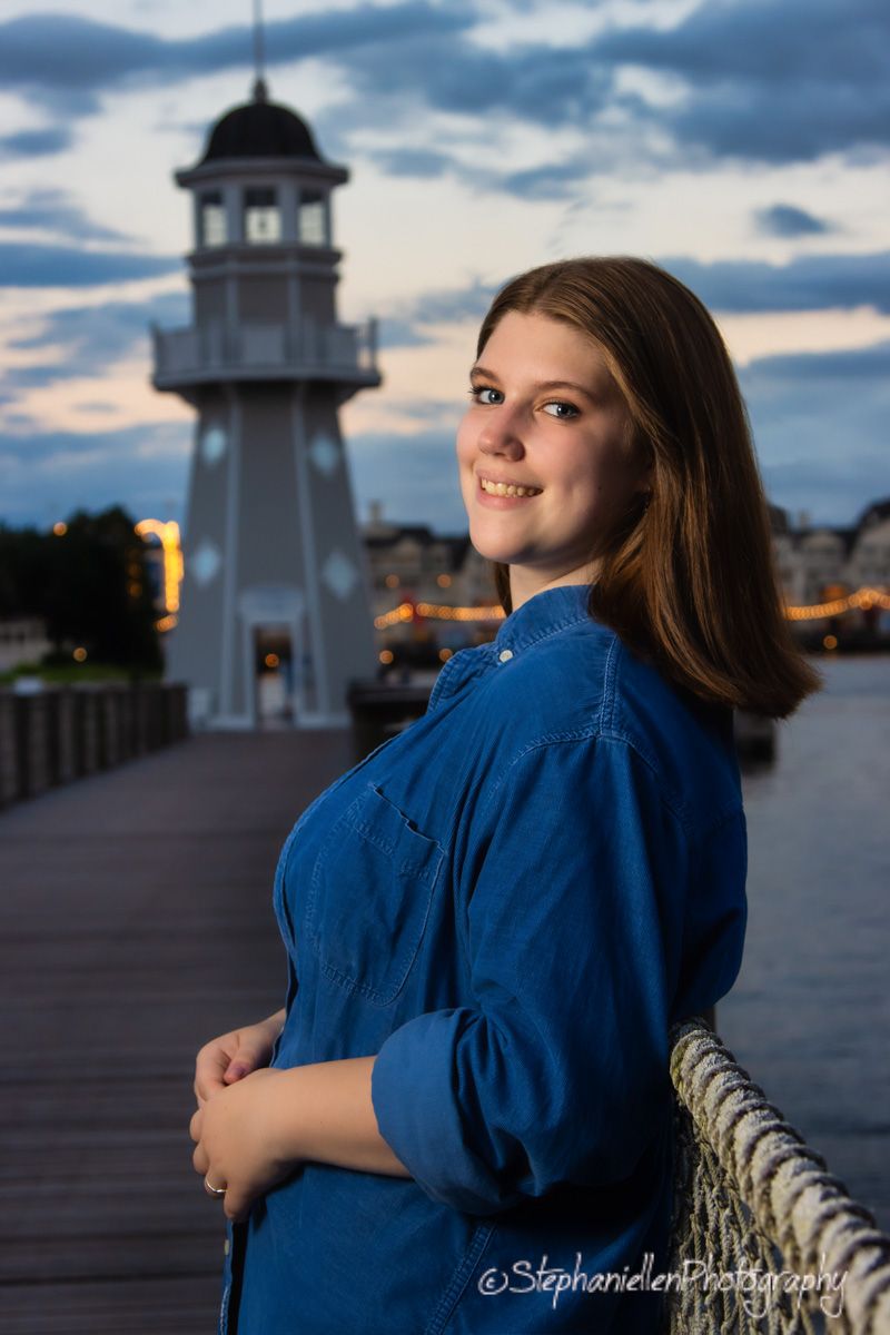 Smiling young woman in blue denim shirt posing near lighthouse at dusk waterfront.