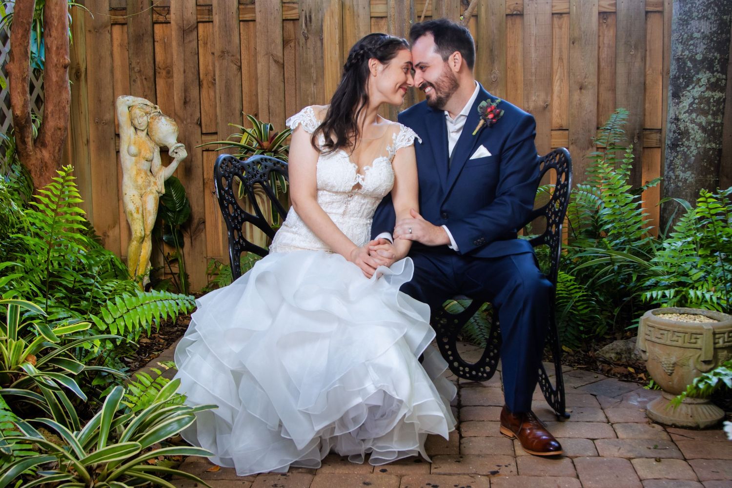 Bride and groom sit closely on ornate bench in lush garden, sharing a tender smile on their wedding day.