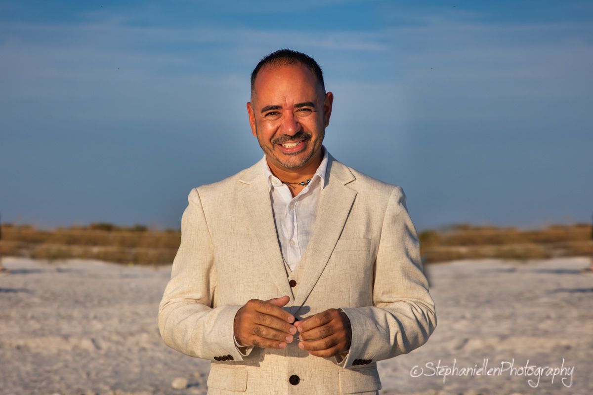 Smiling man in a beige suit posing on a sunny beach with blue sky.