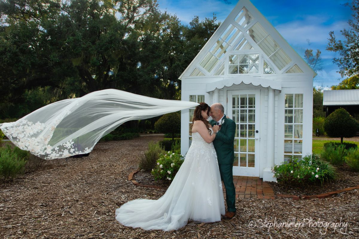 A bride and groom share a kiss as her dramatic veil billows in the wind beside a charming garden chapel.