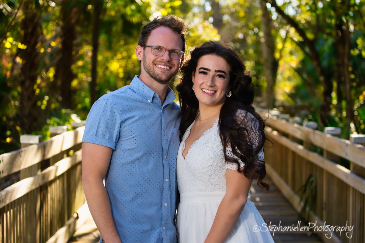 A smiling couple poses on a wooden bridge surrounded by lush greenery in a professional outdoor portrait photo.